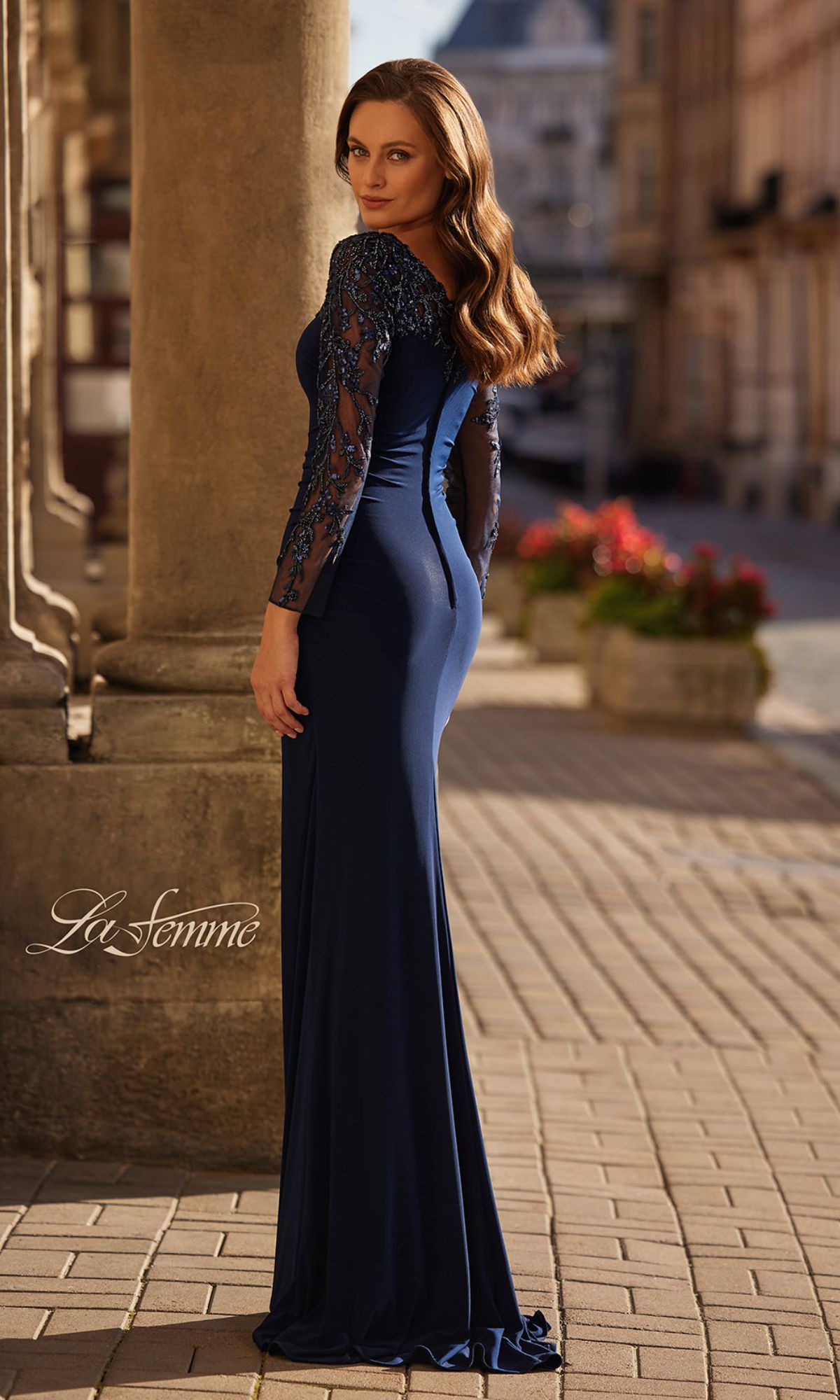 A woman stands outdoors on a cobblestone street, looking over her shoulder in the fitted Long Sleeve Long Navy Formal Dress: La Femme 32609, featuring elegant lace sleeves.